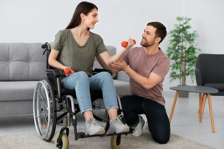 Eine junge Frau im Rollstuhl trainiert lachend gemeinsam mit einem Begleiter im Wohnzimmer.