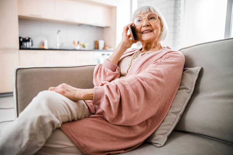 Eine ältere Dame sitzt entspannt auf dem Sofa und telefoniert lächelnd mit ihrem Smartphone.